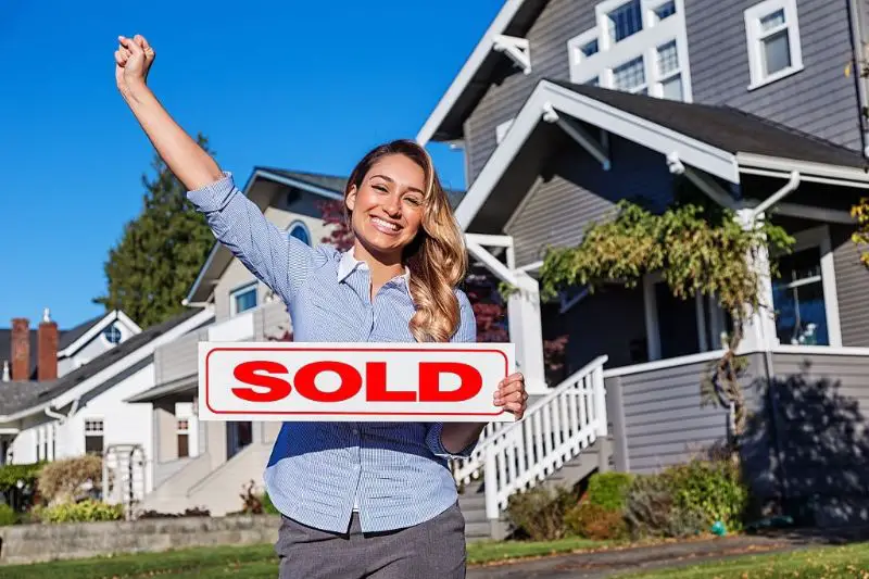 a cheerful young women with a red SOLD sign in front of a row of houses